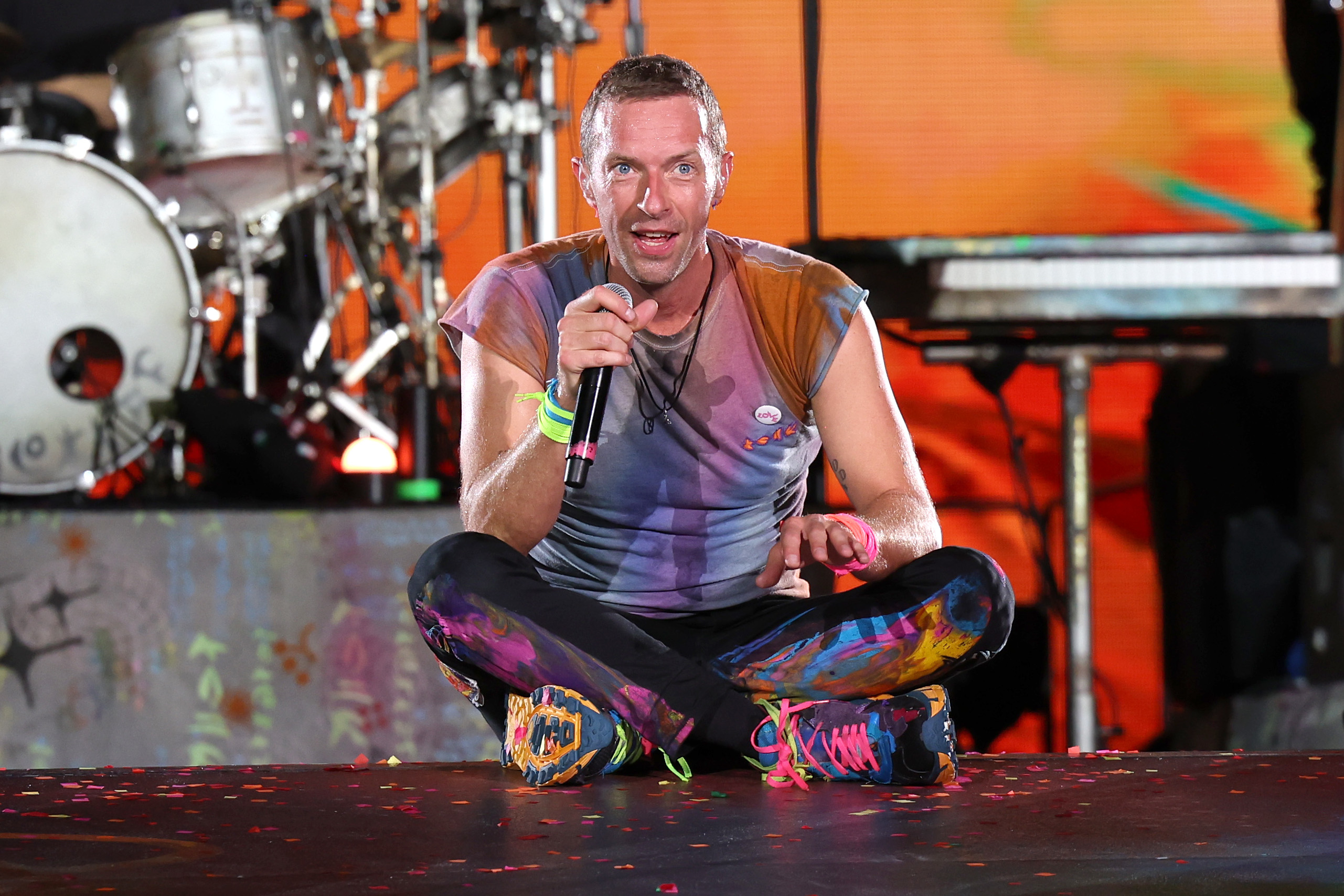 Chris Martin, Coldplay's lead vocalist, sits on stage during a performance, holding a microphone and wearing vibrant, colorful clothing