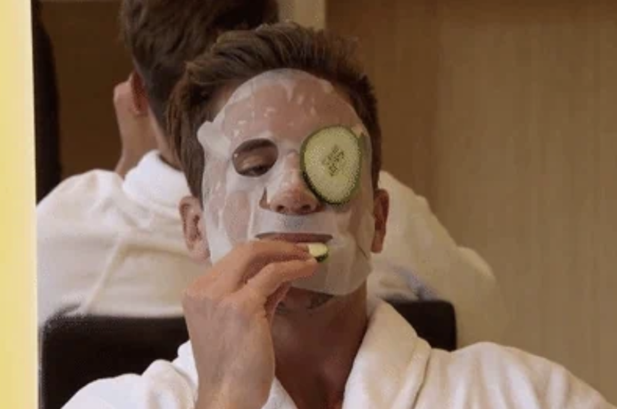 Man wearing a white robe, face mask, and a cucumber slice on one eye, while eating a cucumber slice