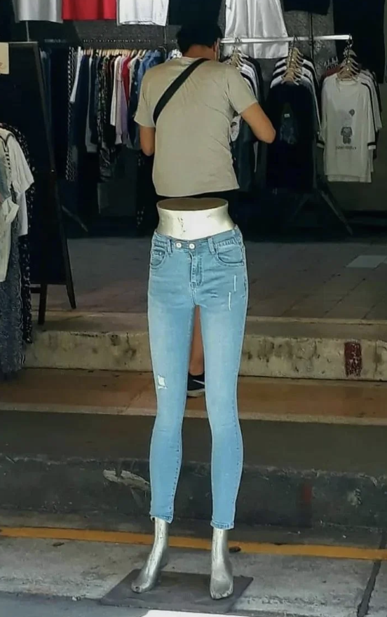 A person adjusting clothes on a hanger in a store, seen from behind a mannequin displaying jeans and silver shoes. The mannequin's upper body is missing