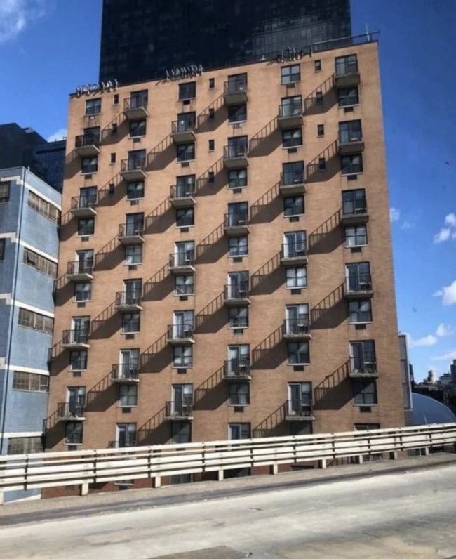 A tall brick building with repeating balconies and fire escapes, viewed from an angle. Nearby are other buildings and a sky with some clouds