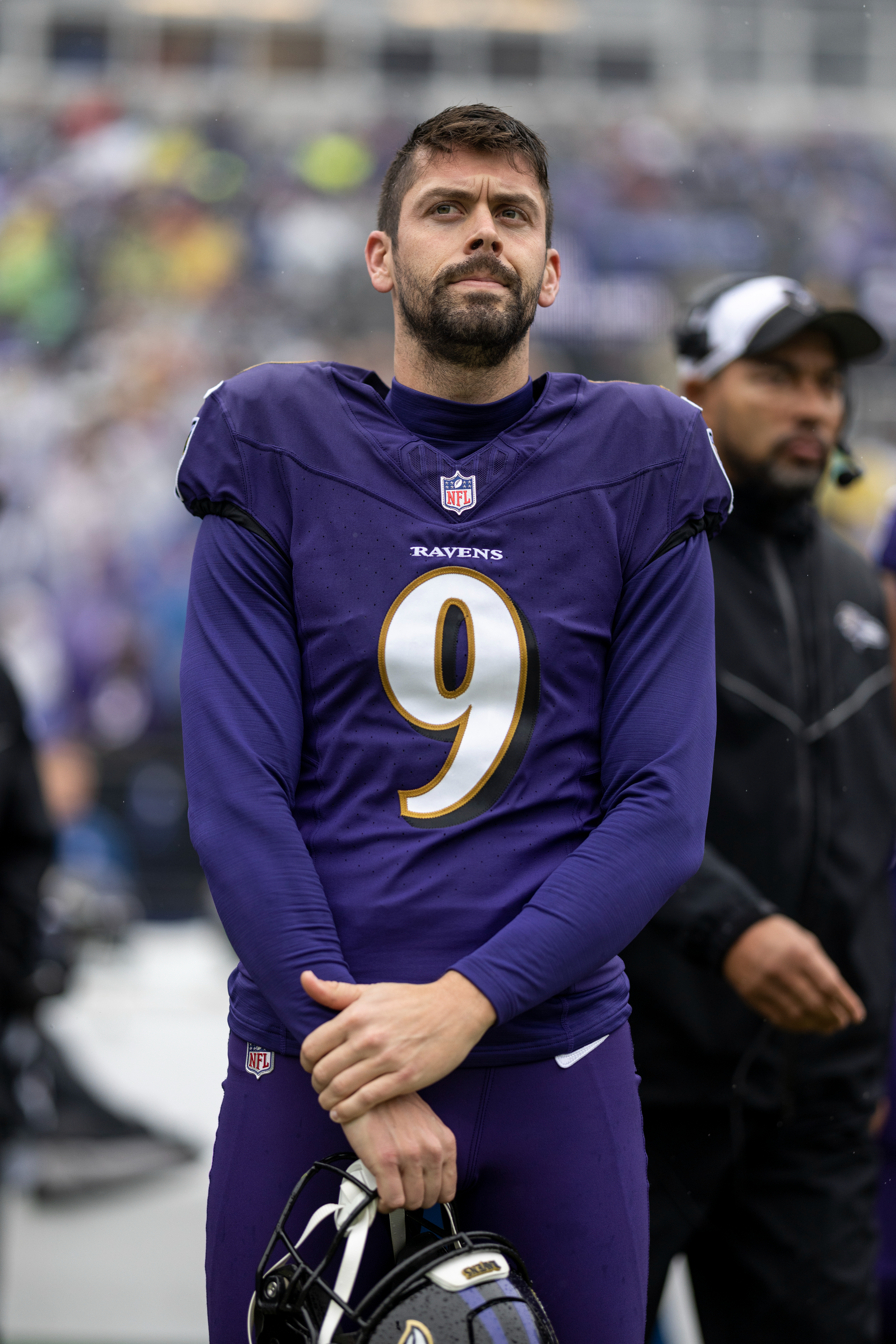 Justin Tucker in a Ravens football uniform on the field, holding his helmet and looking contemplative. A team staff member stands in the background