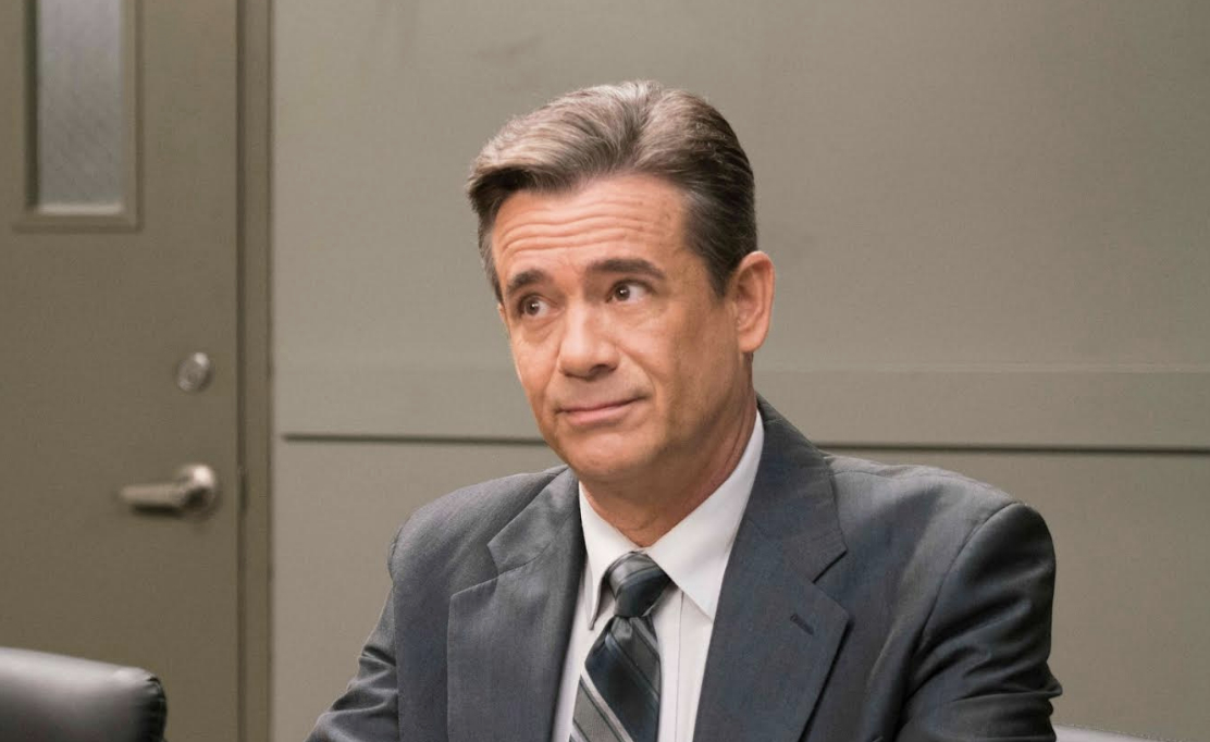 Daran Norris, wearing a suit and tie, sits at a table looking slightly amused