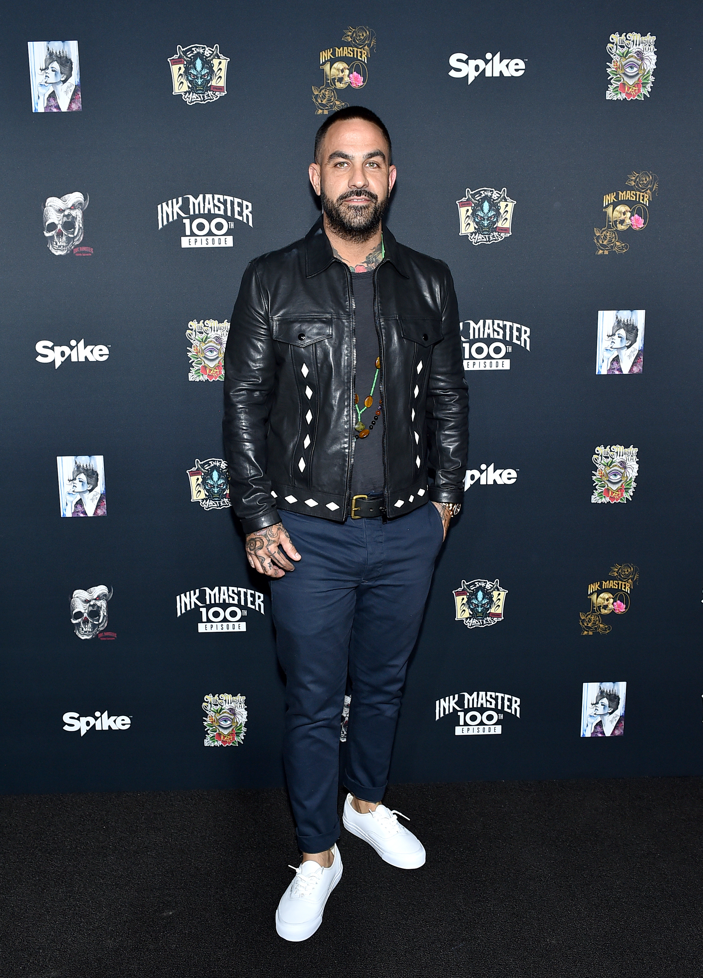 Chris Nunez on the red carpet, wearing a black leather jacket, dark shirt, blue pants, and white shoes, at an event for Spike's Ink Master 100th episode celebration