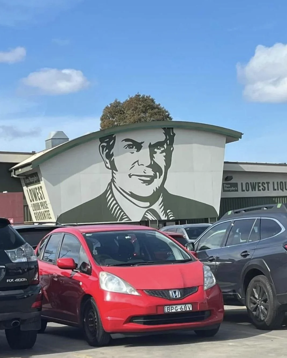 Large mural of a man's face painted on the side of a building in a parking lot. The building signage reads "Lowest Liquor & Cigarette" and "Northern Hotel"