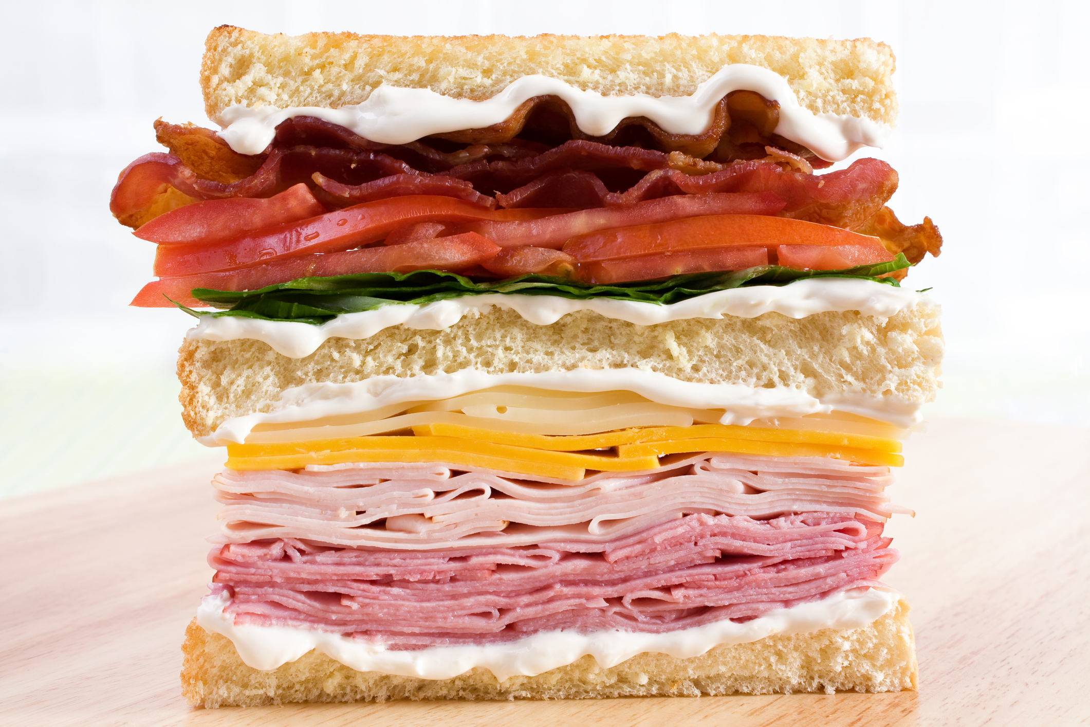 Stacked sandwich with various layers including bacon, tomato, lettuce, cheese, turkey, and pastrami between slices of bread