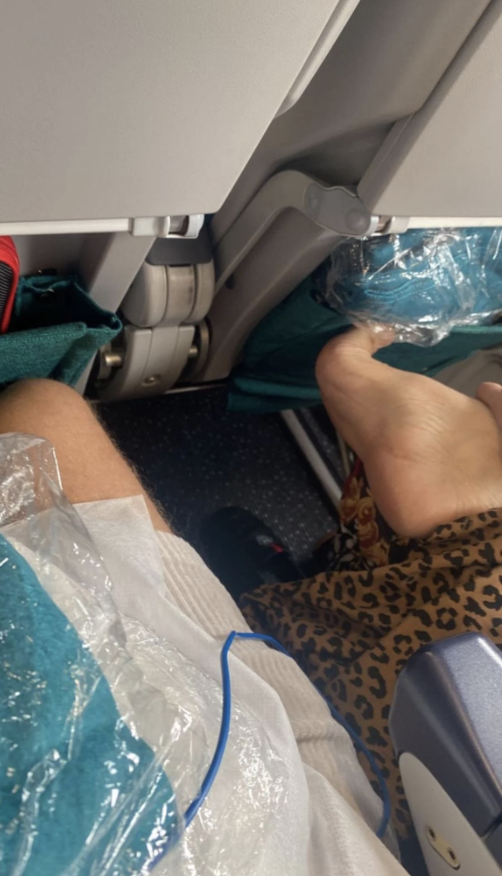 A person's legs, partly covered, rest on an airplane tray table with a partial view of another passenger's feet wrapped in plastic