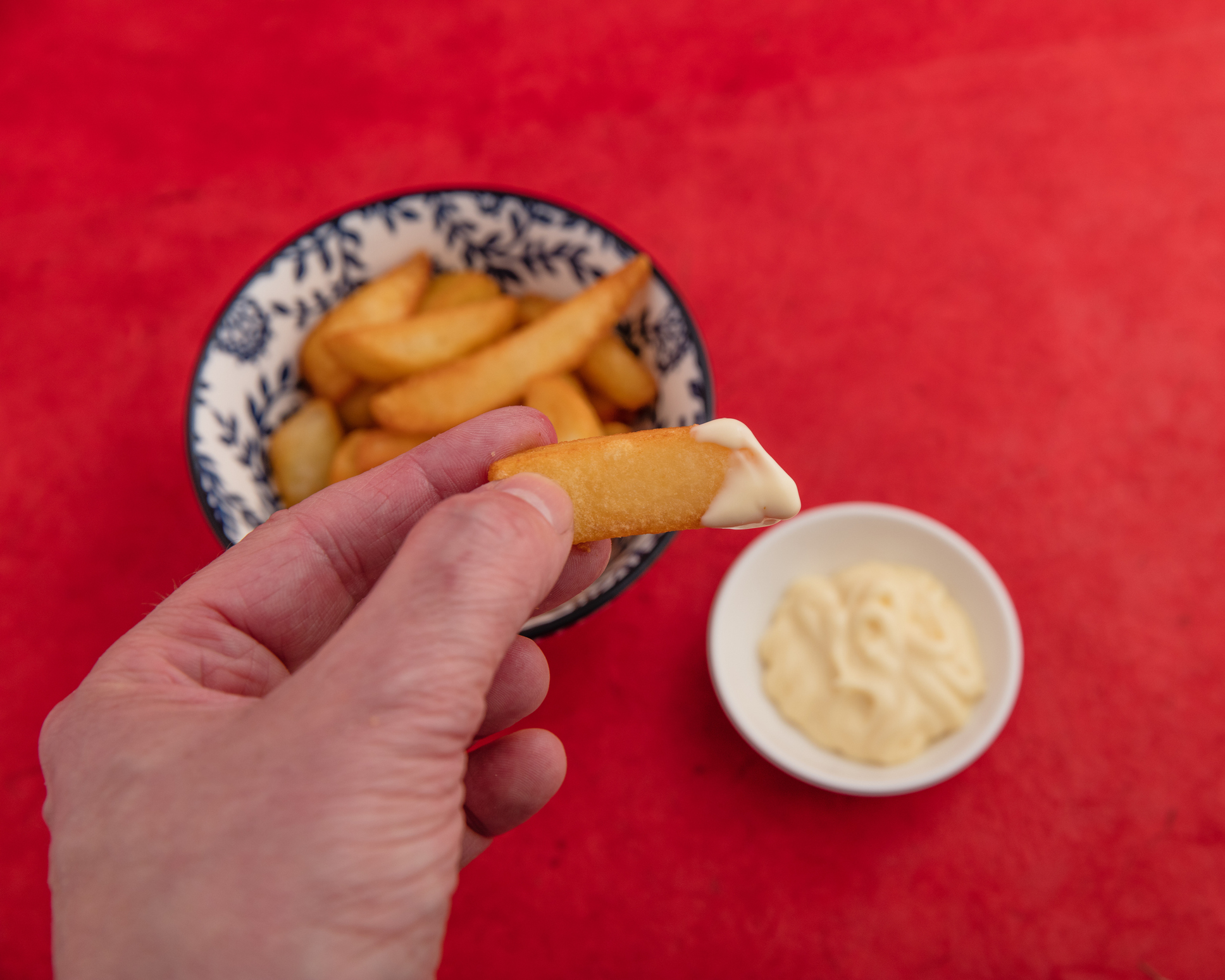 A hand holding a chip with mayonnaise over a bowl of more chips, and an additional small bowl of mayonnaise on the side