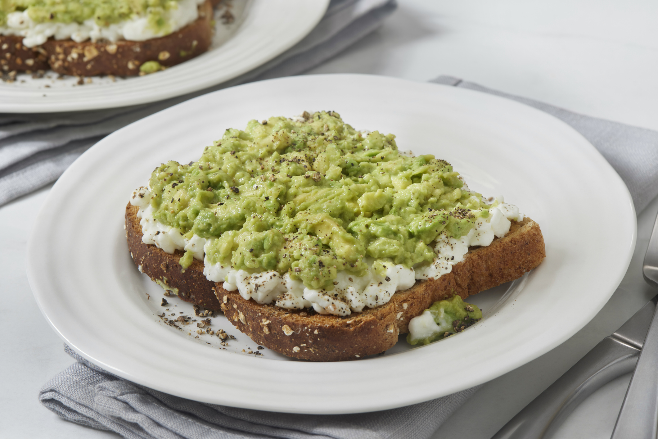 A plate with avocado toast topped with crumbled cheese and seasonings on a white tablecloth. Another plate with avocado toast is blurred in the background
