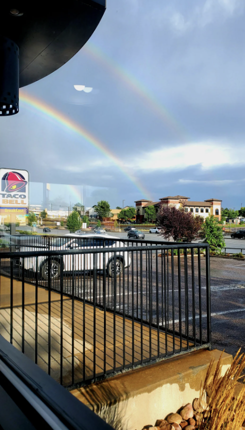A Taco Bell is visible through a window with a double rainbow in the background over the parking lot and surrounding buildings. There are no people in the image