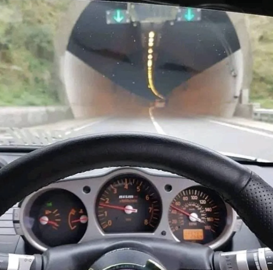 View from inside a vehicle, dashboard and steering wheel visible, approaching a tunnel with electronic signs indicating lanes with green arrows