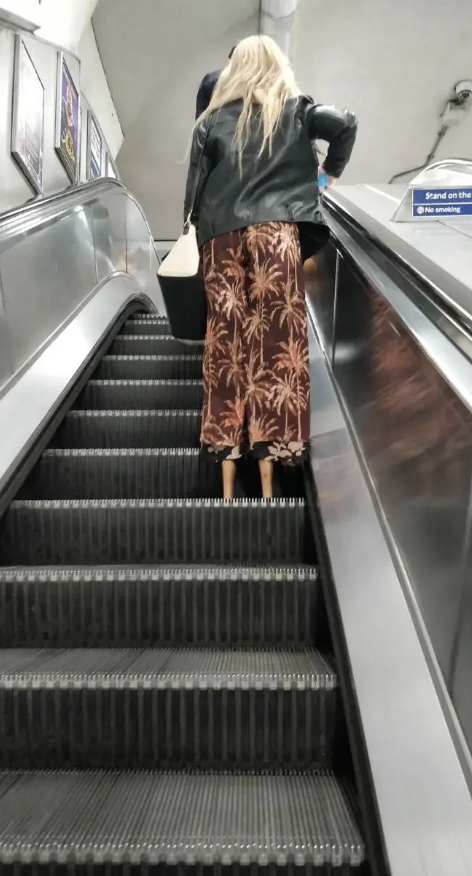 Person with long hair in a leather jacket and patterned skirt riding up an escalator in a subway station