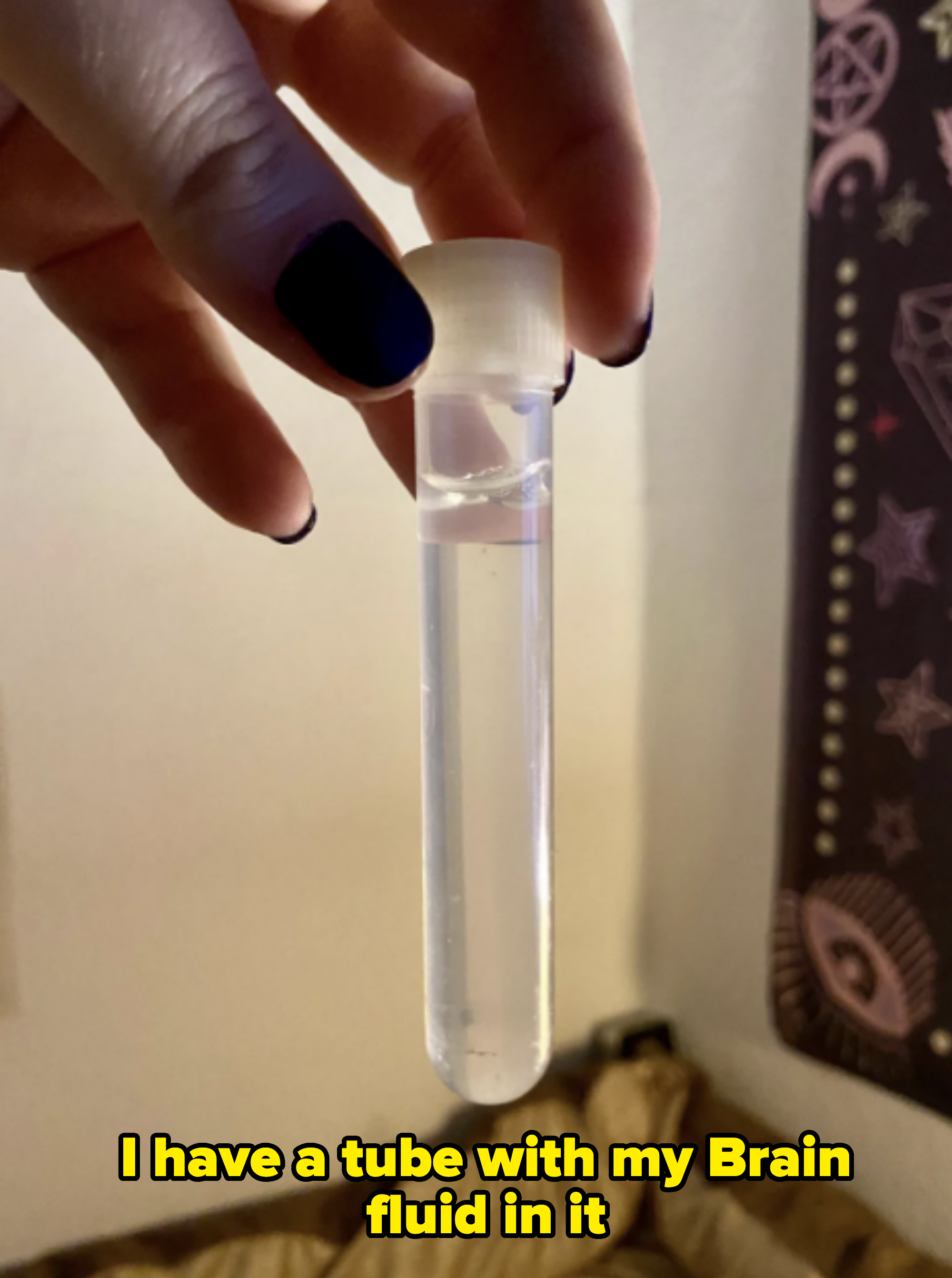 A hand with black-painted nails holds a small clear vial filled with liquid against a blurred background with decorative elements