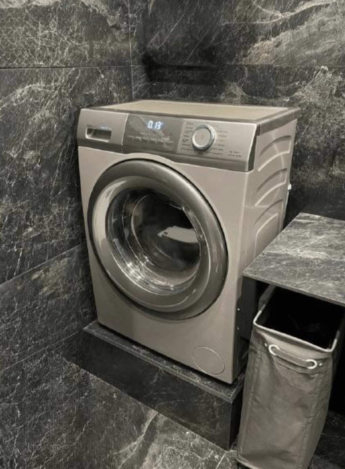 Modern front-loading washing machine in a tiled laundry room, next to a laundry hamper