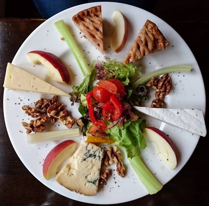 A round plate with assorted cheeses, apple slices, celery sticks, red pepper, nuts, and a leafy green salad in the center