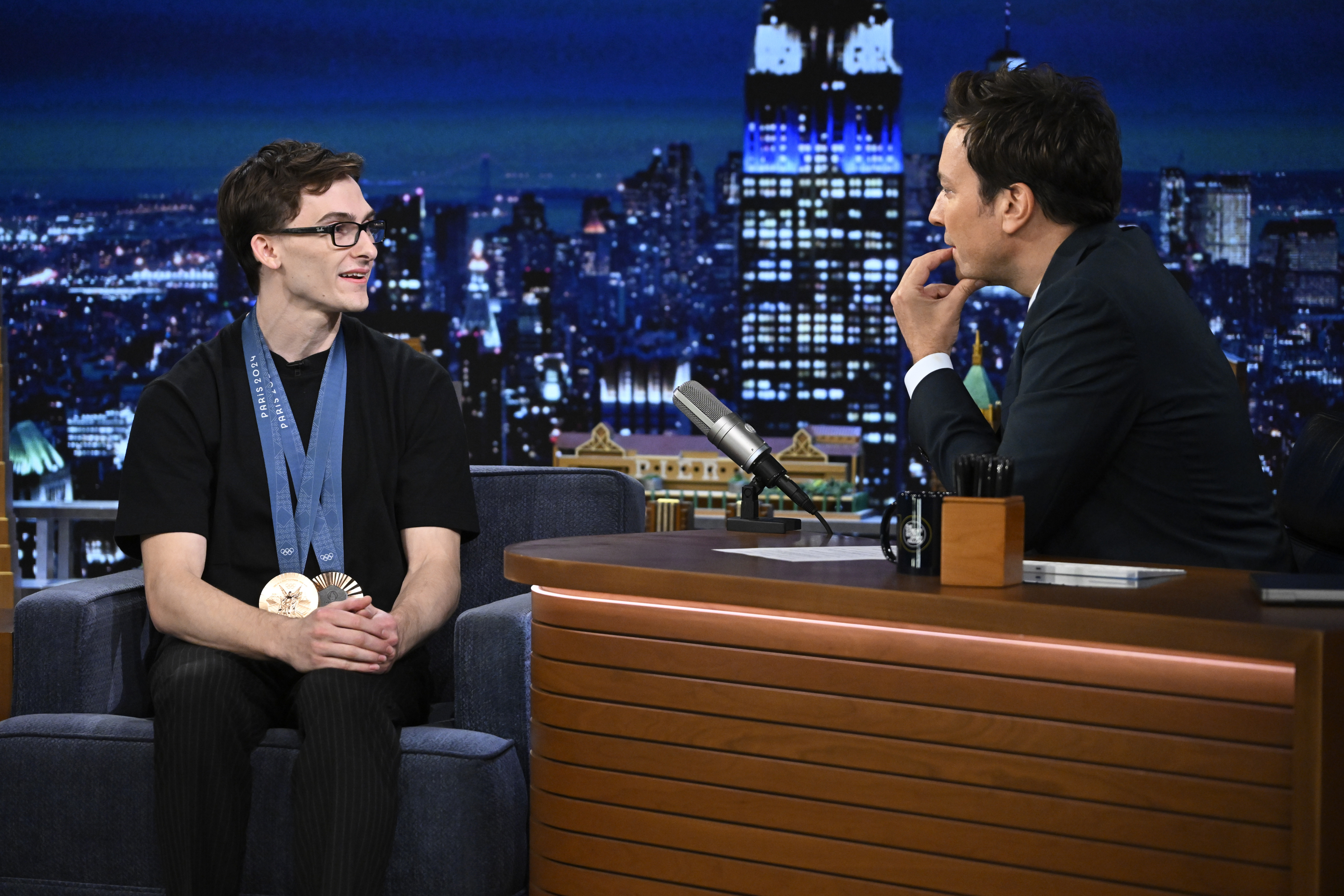 Anthony Veer sits on a talk show couch with multiple medals around his neck, conversing with host Jimmy Fallon at a desk. A cityscape of New York City is in the background