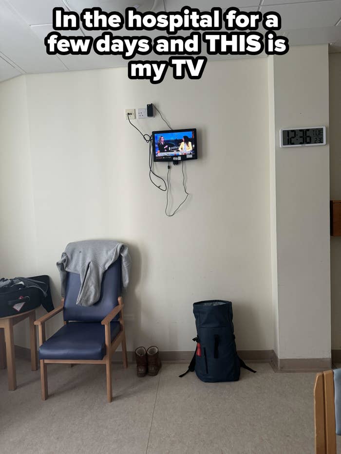 Hospital room with a chair, clothes draped on it, bags on the floor, a small TV mounted on the wall showing a talk show, and a wall clock reading 12:36
