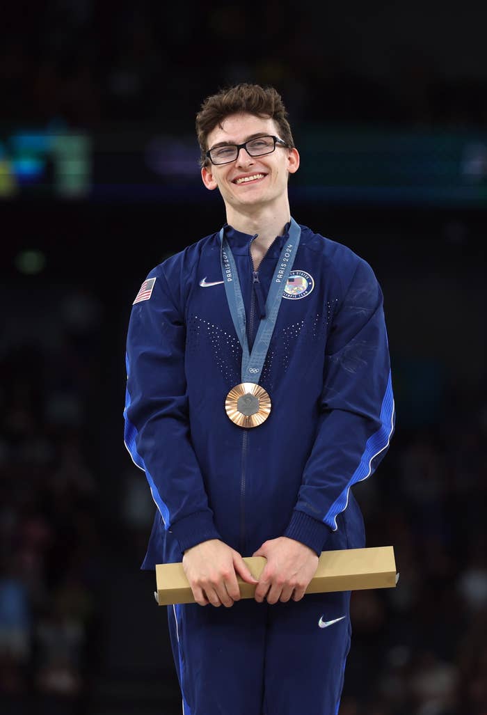 Stephen Nedoroscik wearing a team USA tracksuit and glasses stands smiling with a bronze medal and a wooden plaque in hand