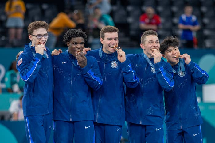 Team USA male gymnasts celebrate, biting gold medals, in matching blue athletic wear on a podium