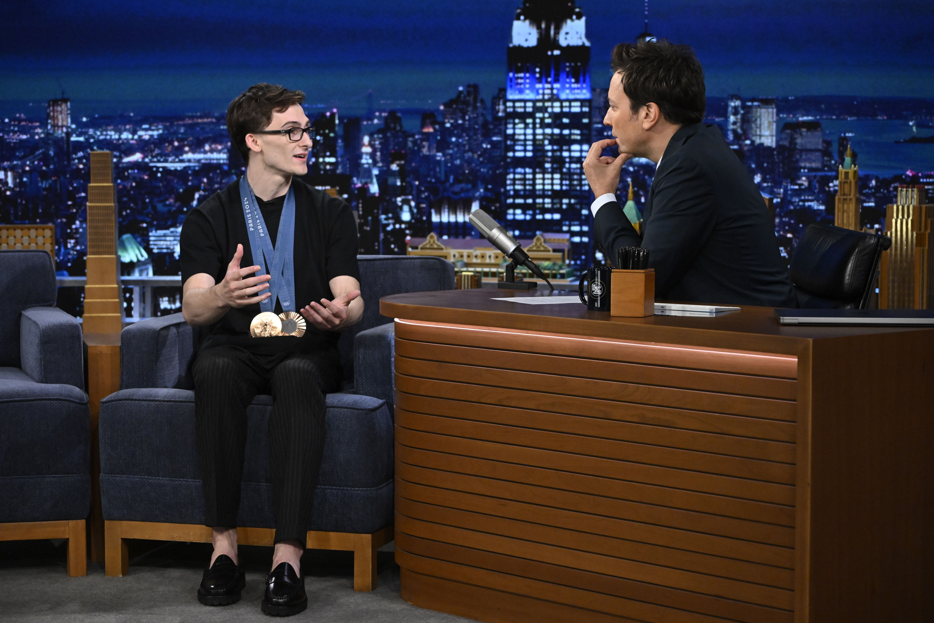 Tom Holland talks with Jimmy Fallon on &quot;The Tonight Show&quot; set, with a New York City skyline background. Tom wears a casual outfit and glasses, Jimmy in a suit