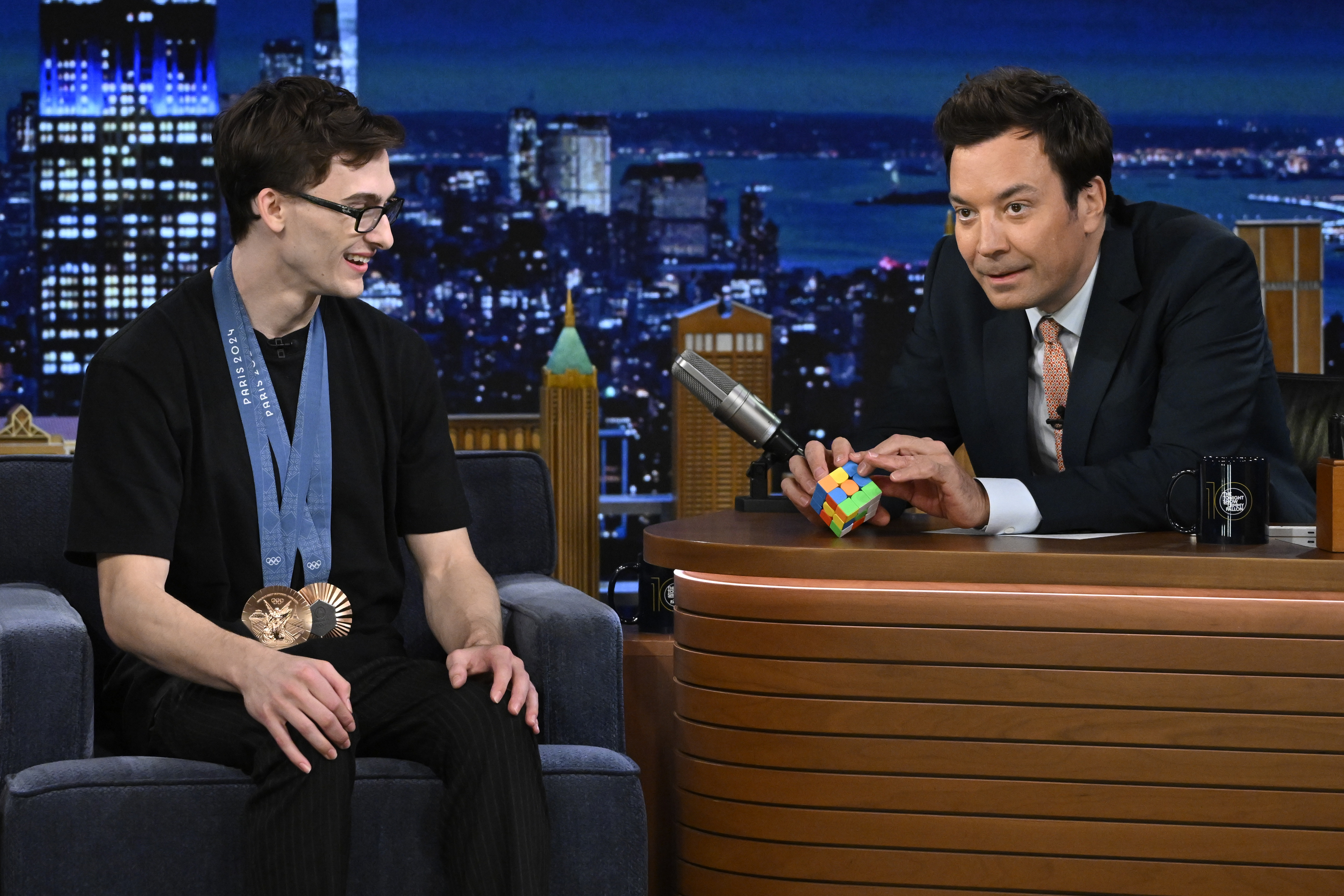 Stephen Nedoroscik, wearing a medal, sits on a talk show set while Jimmy Fallon holds a Rubik's Cube during an interview