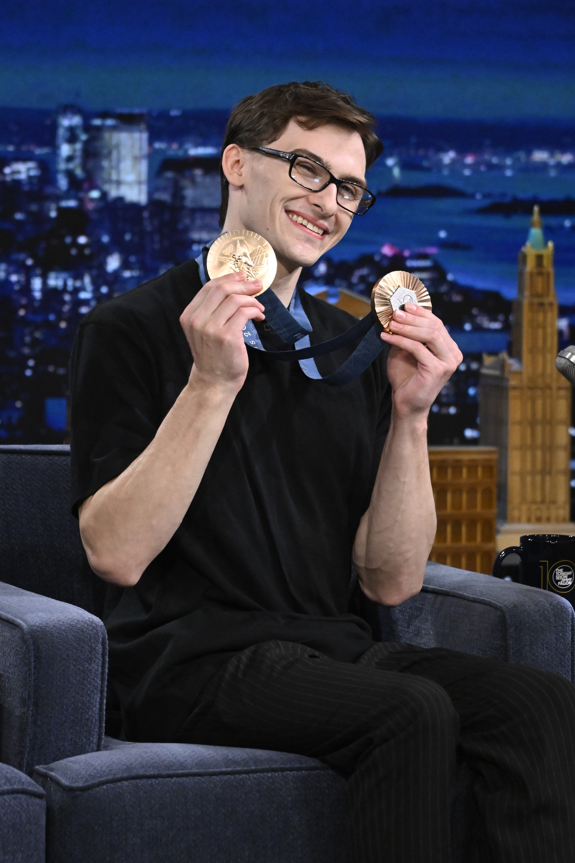 Man sitting on a talk show set, smiling and holding two medals. He is wearing glasses and casual black clothing