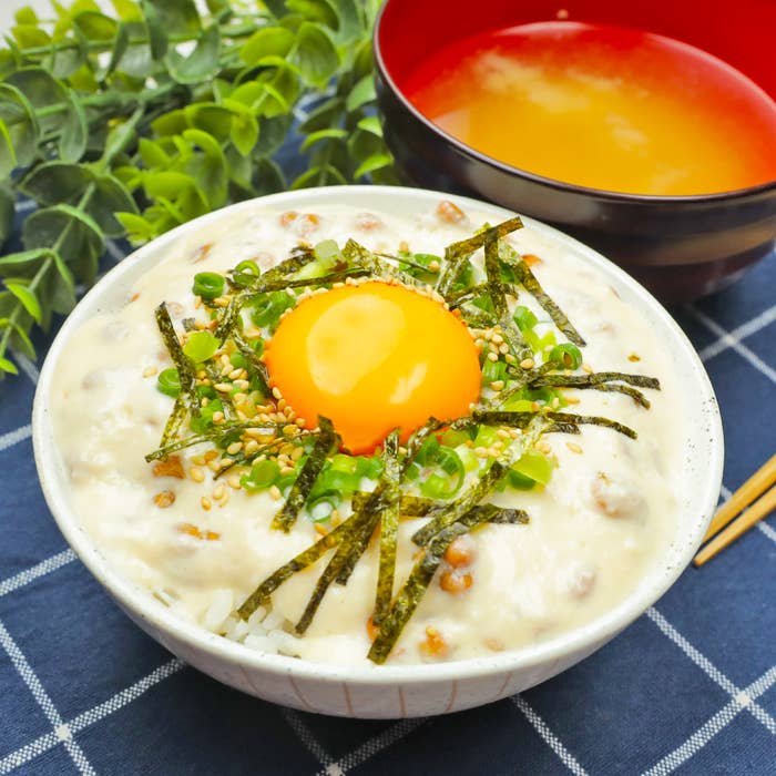 ご飯の上に納豆、刻んだ海苔、青ネギ、生卵の黄身が乗った白い器の料理と、味噌汁が並んでいる。