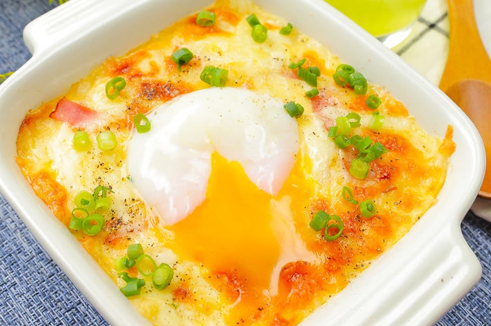 白い器に盛られたグラタン風料理。中央に半熟の目玉焼きがトッピングされ、ネギが散らされています。背景には黄色いドリンクと花が見えます。