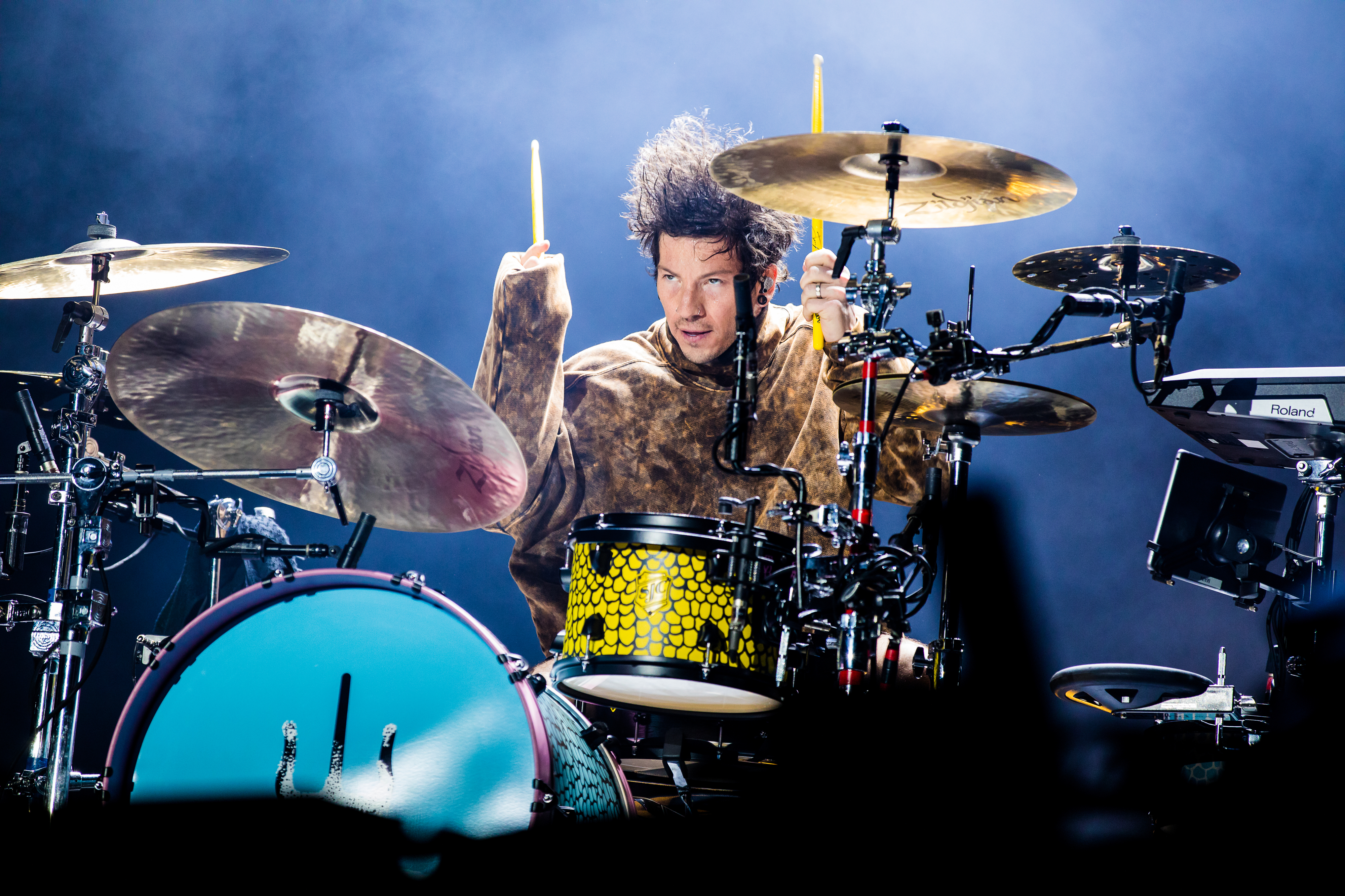Josh Dun playing drums energetically on stage, surrounded by a full drum kit and lighting equipment
