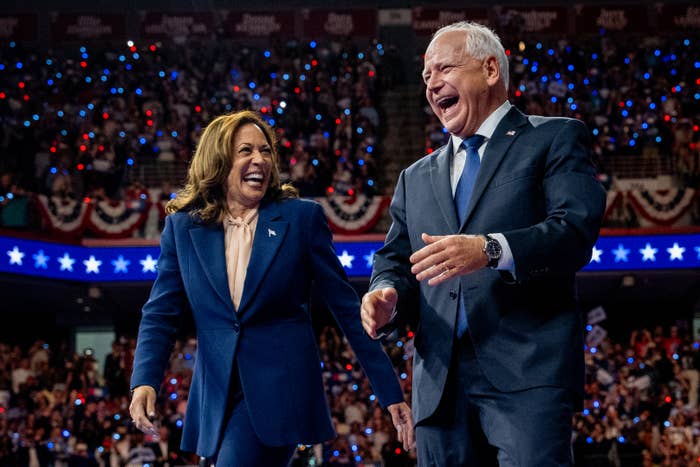 Kamala Harris and Tim Walz, both wearing suits, share a laugh on stage at a crowded event