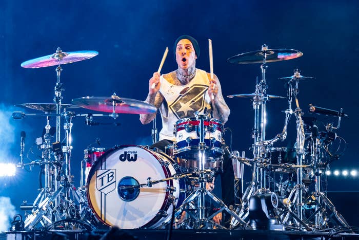 Travis Barker plays the drums on stage during a live performance. He is wearing a sleeveless top and a beanie. Bright lights and drum equipment surround him