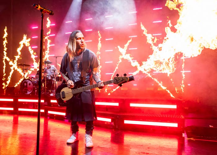 Pete Wentz, holding a bass guitar, performs on stage with dramatic flame effects in the background. A drummer is visible in the background