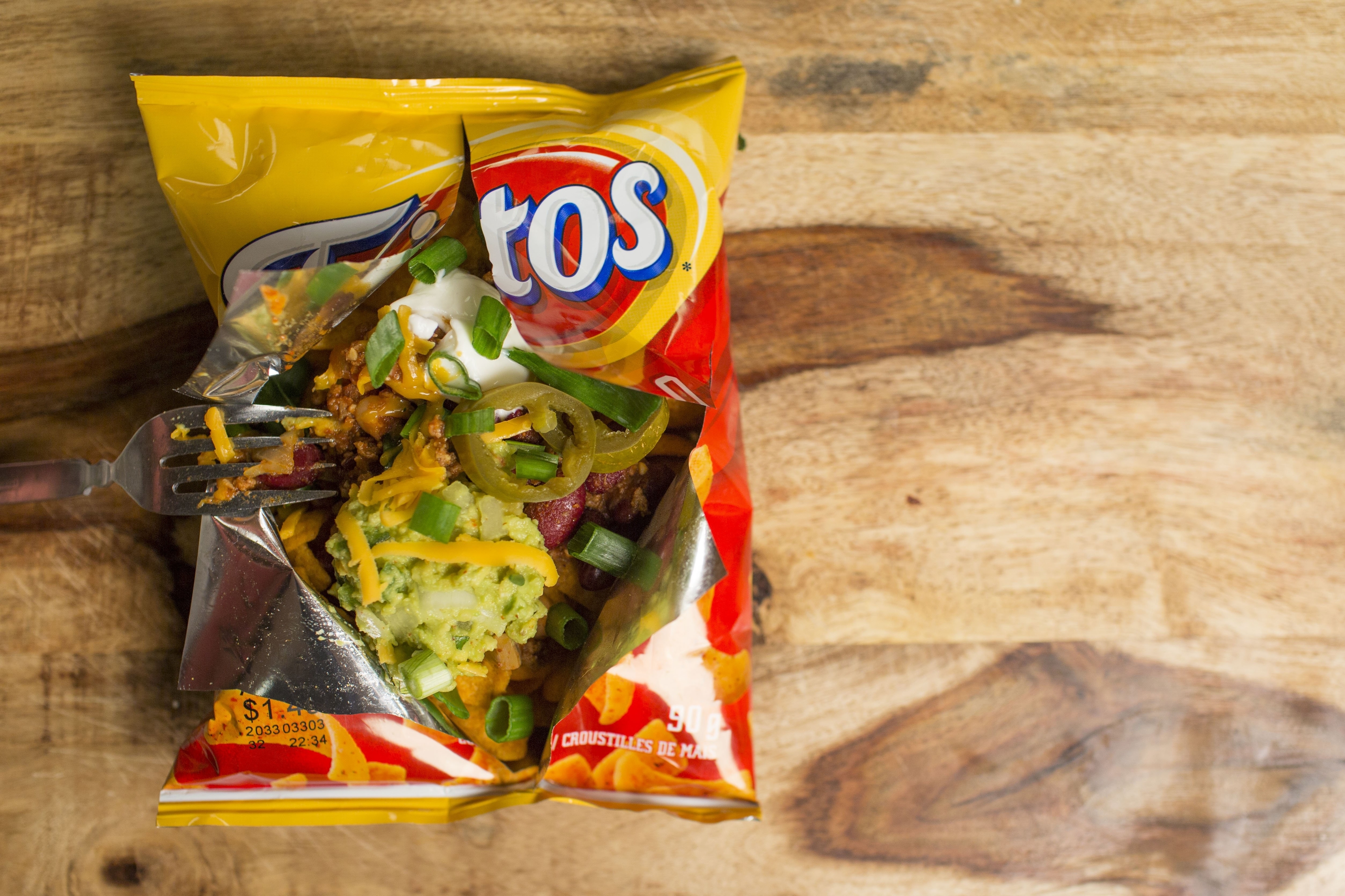 Opened bag of Fritos on a wooden surface, filled with toppings including cheese, green onions, sour cream, guacamole, and jalapeños
