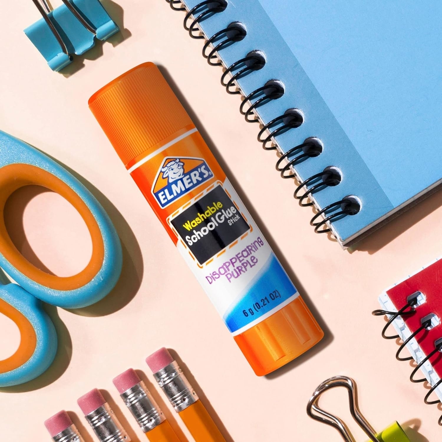 Elmer's washable school glue, scissors, erasers, paper clips, and a spiral notebook are arranged on a surface for a shopping article about back-to-school supplies