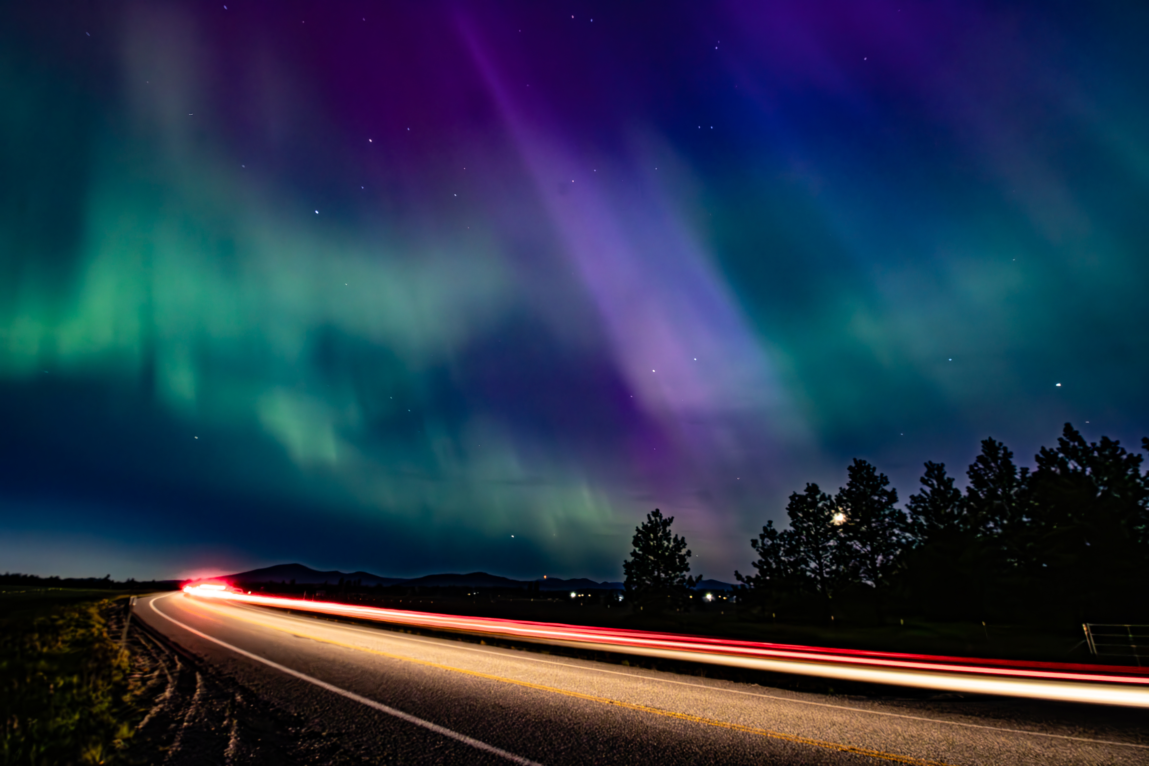 Northern Lights illuminate the night sky over a quiet road with light trails from passing cars, and silhouetted trees in the background