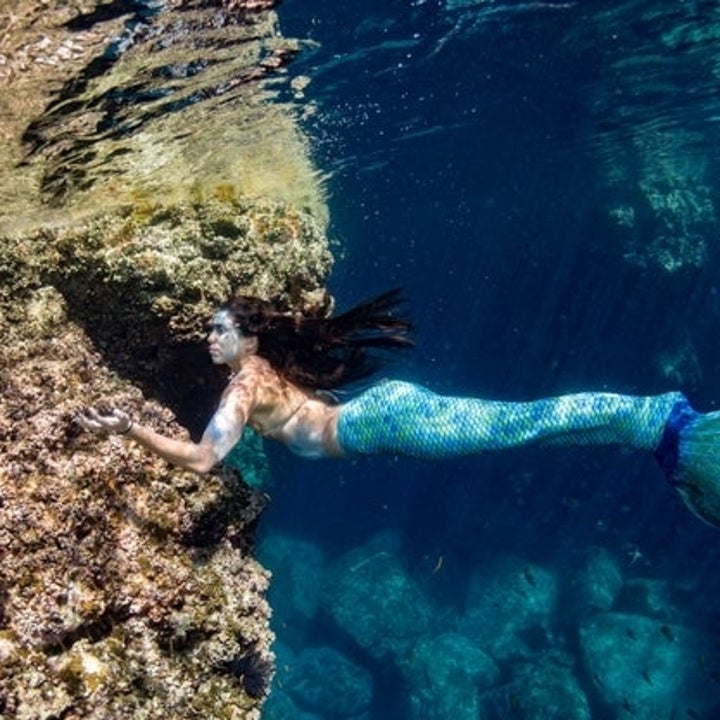 A mermaid is underwater, swimming near rocky terrain. She has long hair and a blue and green tail