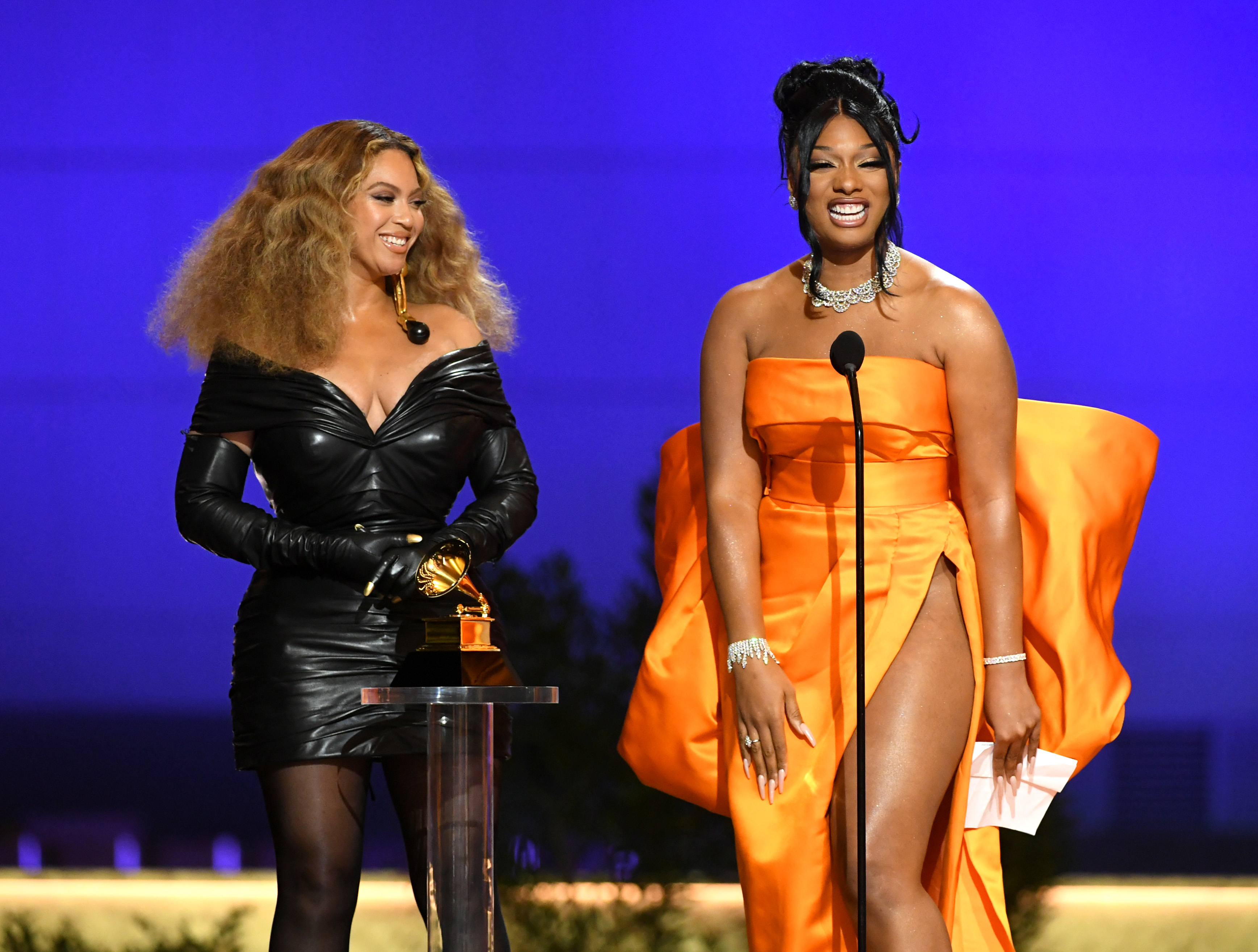 Beyoncé and Megan Thee Stallion on stage at an event, with Beyoncé in an off-shoulder black leather dress and Megan in a strapless orange gown