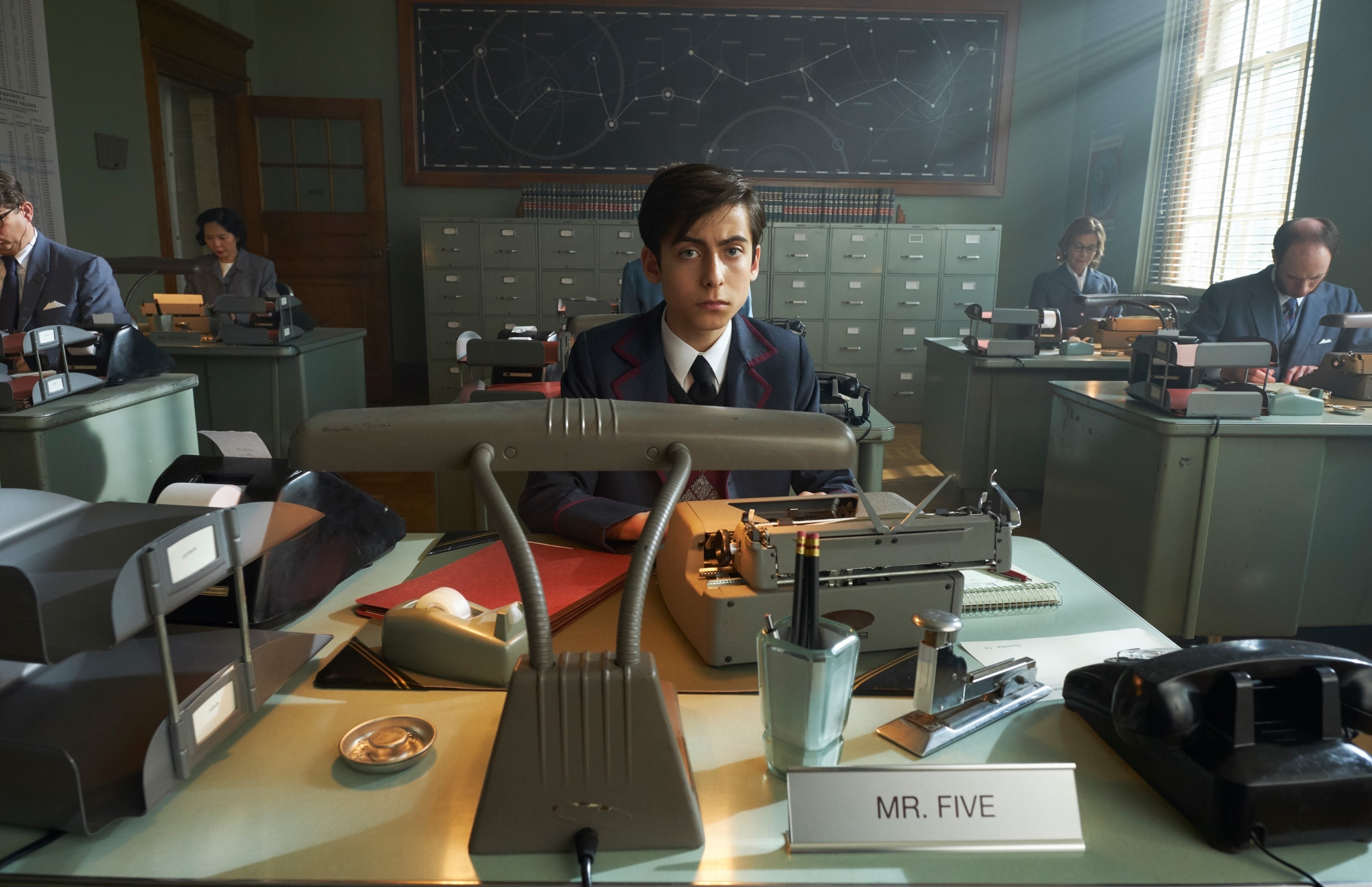 Aiden Gallagher sits at a desk labeled "Mr. Five" in a 1960s-style office filled with colleagues typing on typewriters in a scene from "The Umbrella Academy"