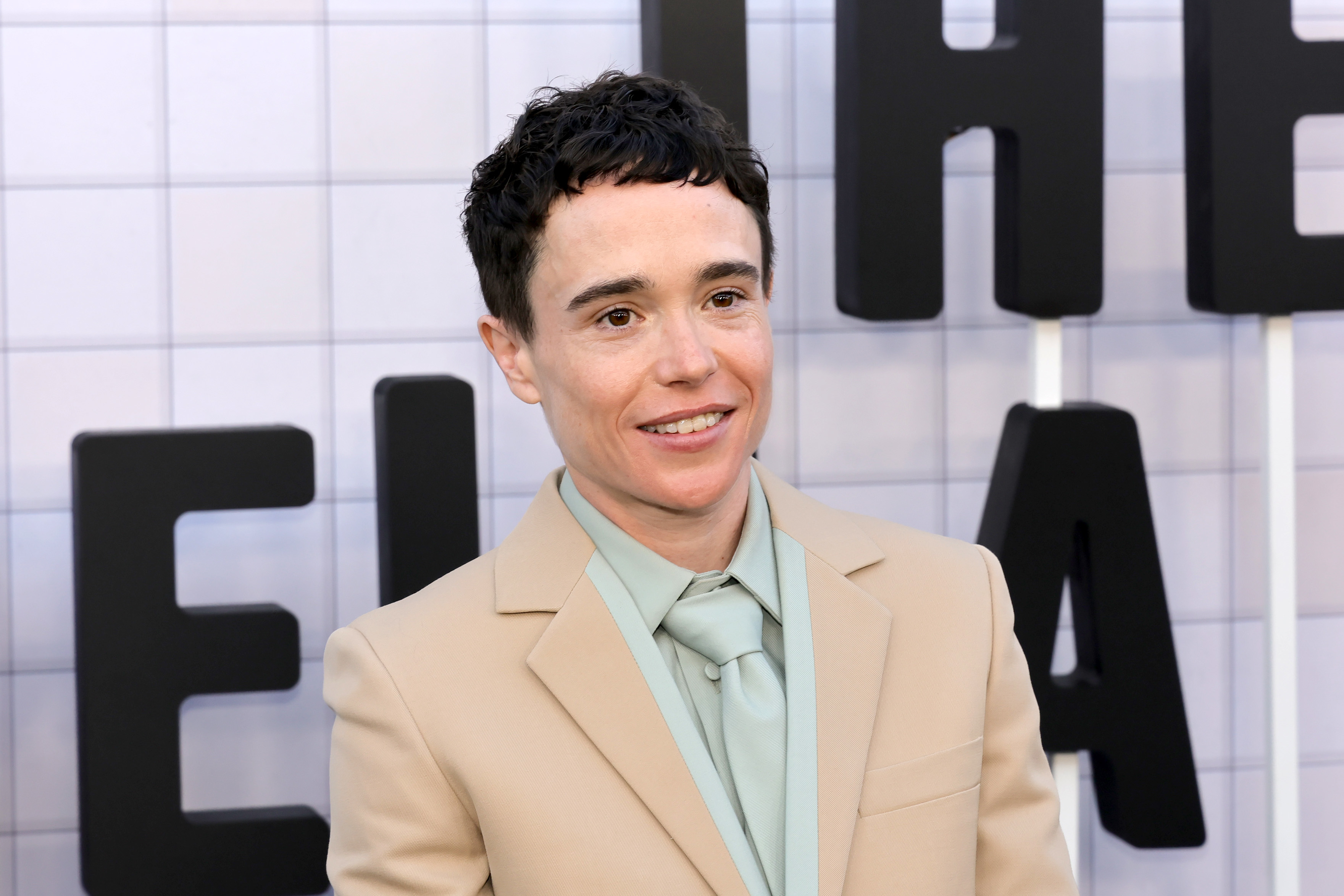 Elliot Page on the red carpet wearing a beige suit with a mint green shirt and tie at a TV event