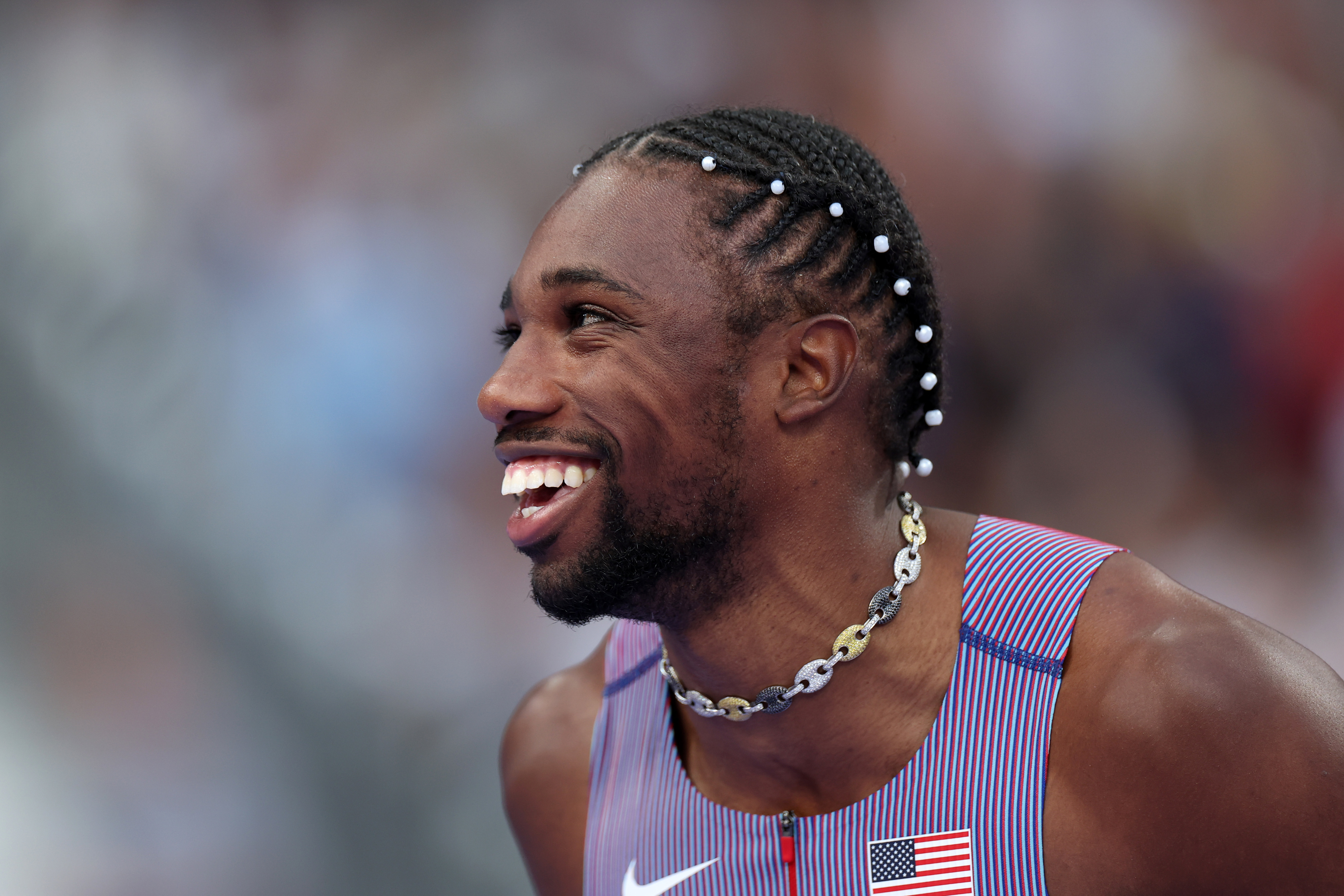A man, wearing a tracksuit adorned with an American flag patch, is smiling widely during what appears to be an athletic event. His hair is styled with braids