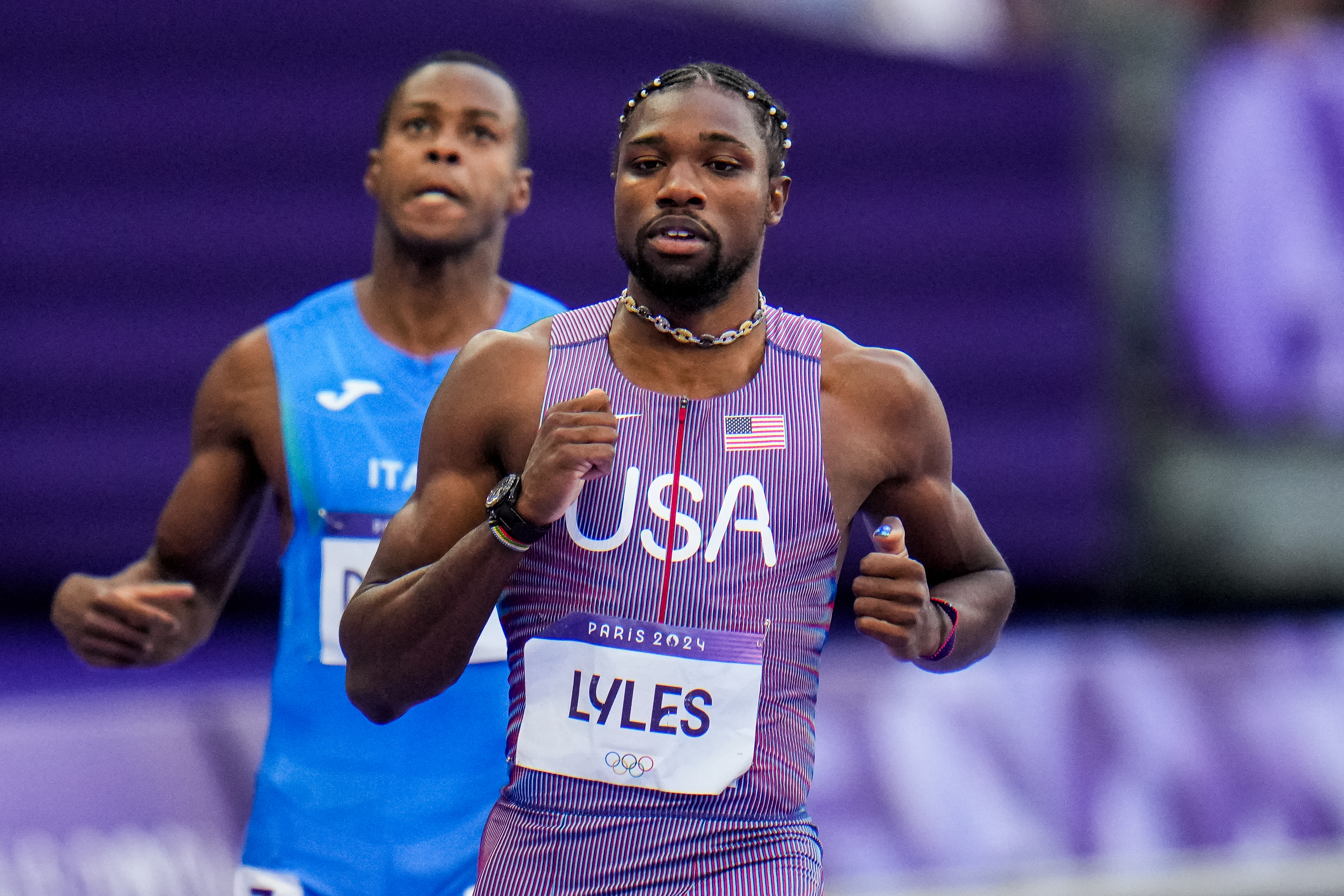 Noah Lyles, wearing a USA uniform, competes in a track event at the Paris 2024 Olympics. Another runner in a blue Italy uniform is visible in the background