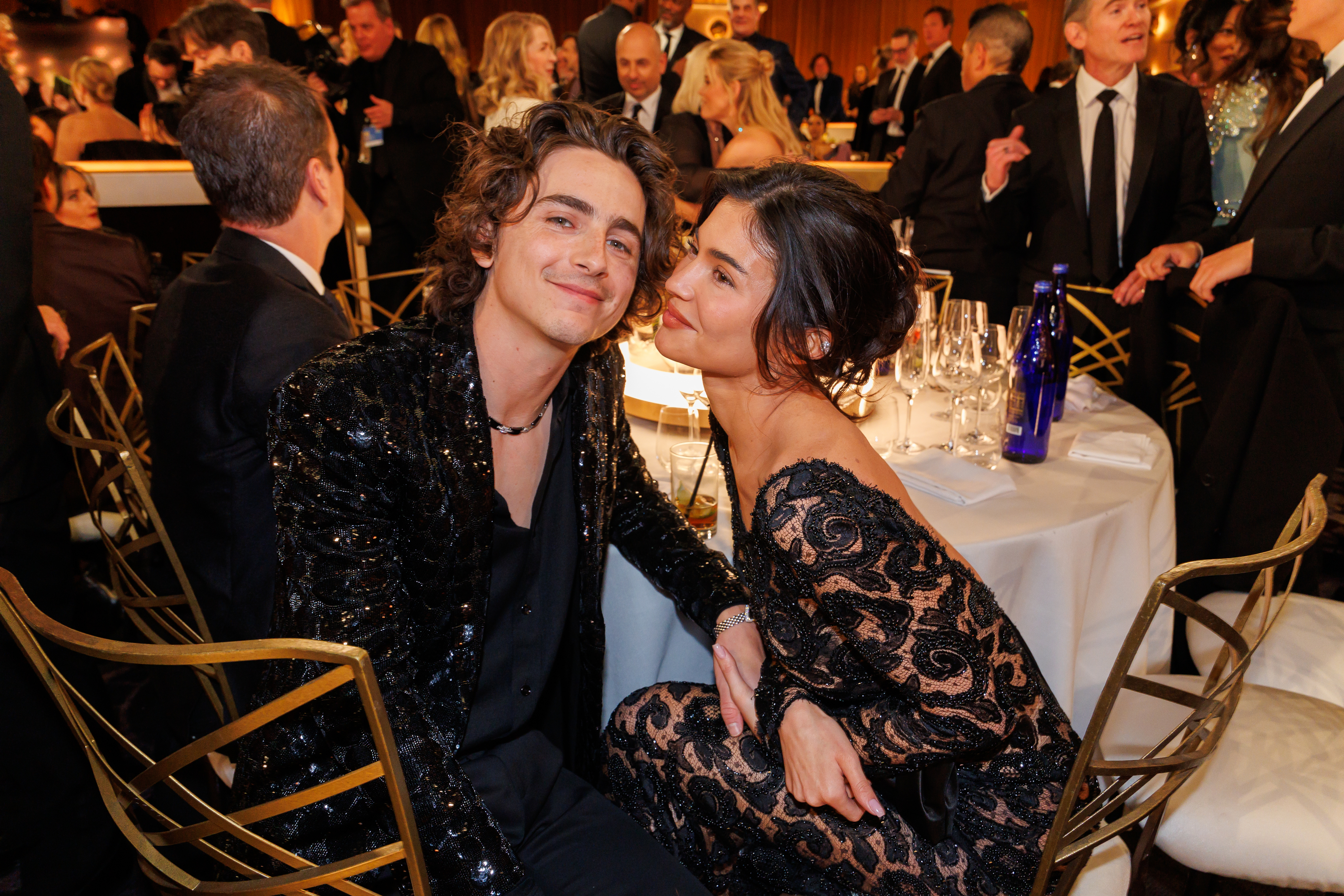 Timothée Chalamet and Kylie Jenner smiling and posing together seated at a formal event. Timothée wears a sequined jacket, and Lily-Rose wears an elegant lace dress