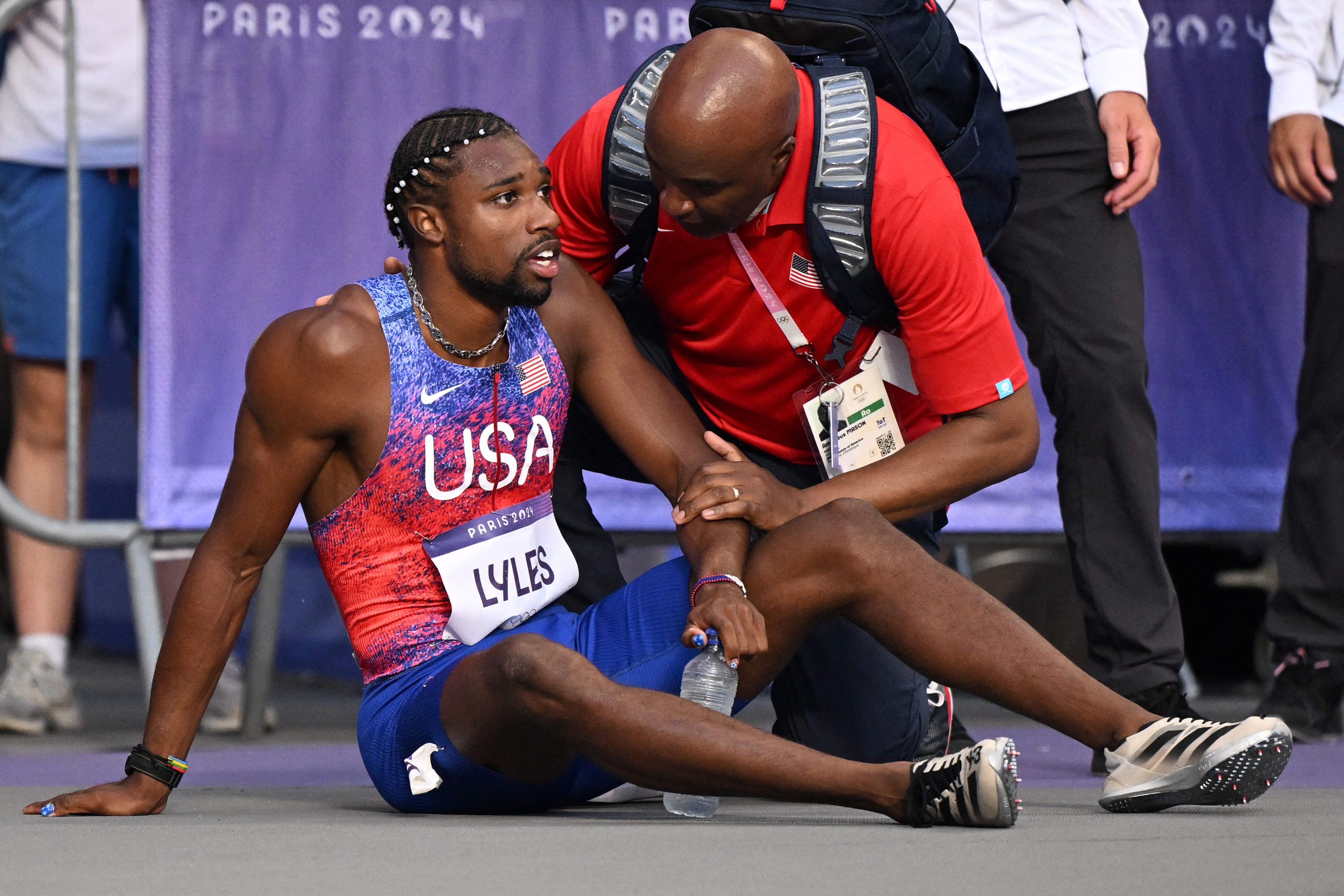 Noah Lyles, wearing a USA athletic uniform, sits on the ground holding a water bottle with a coach or medical personnel assisting him