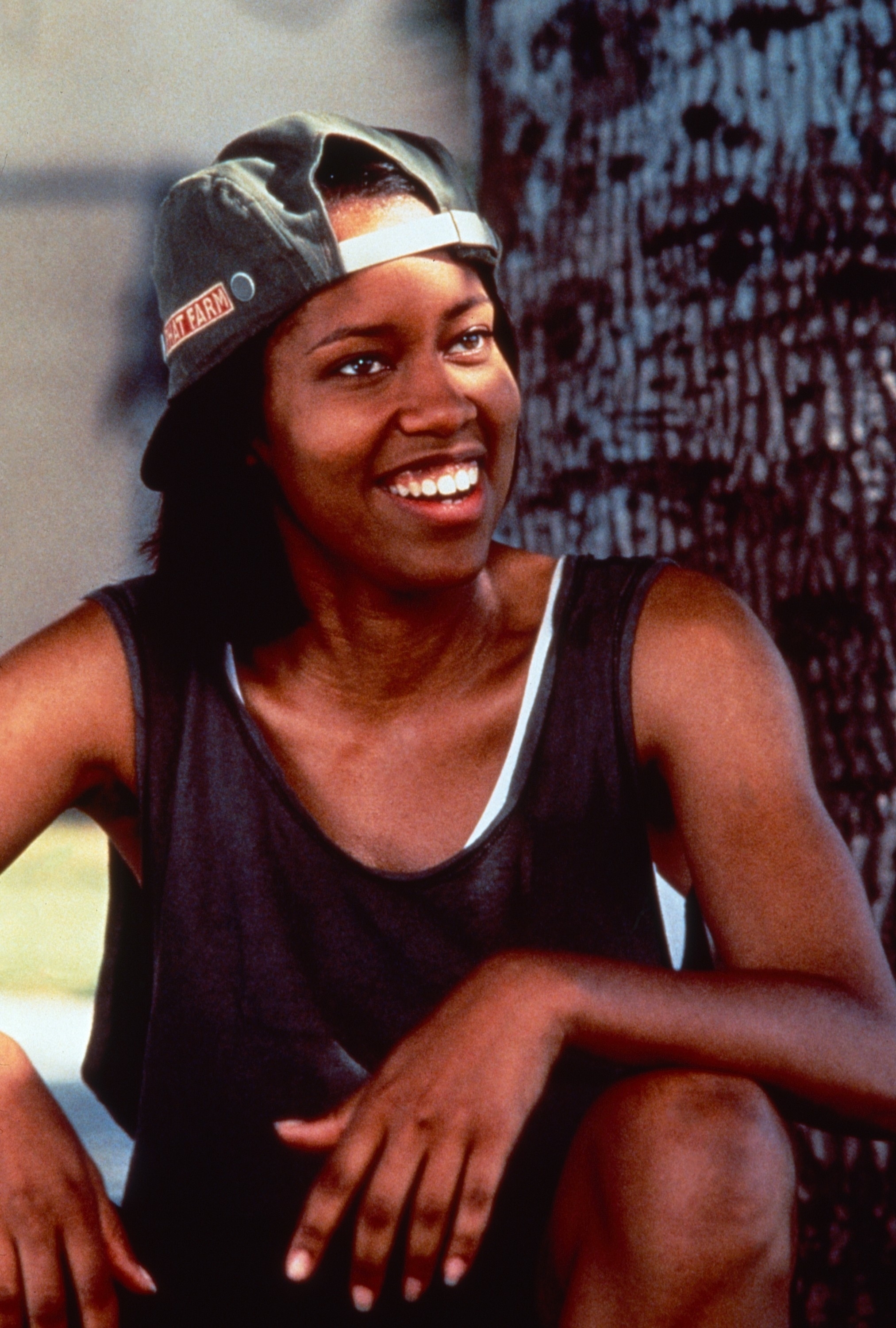 Person sitting outdoors, smiling, wearing a backward cap and sleeveless shirt