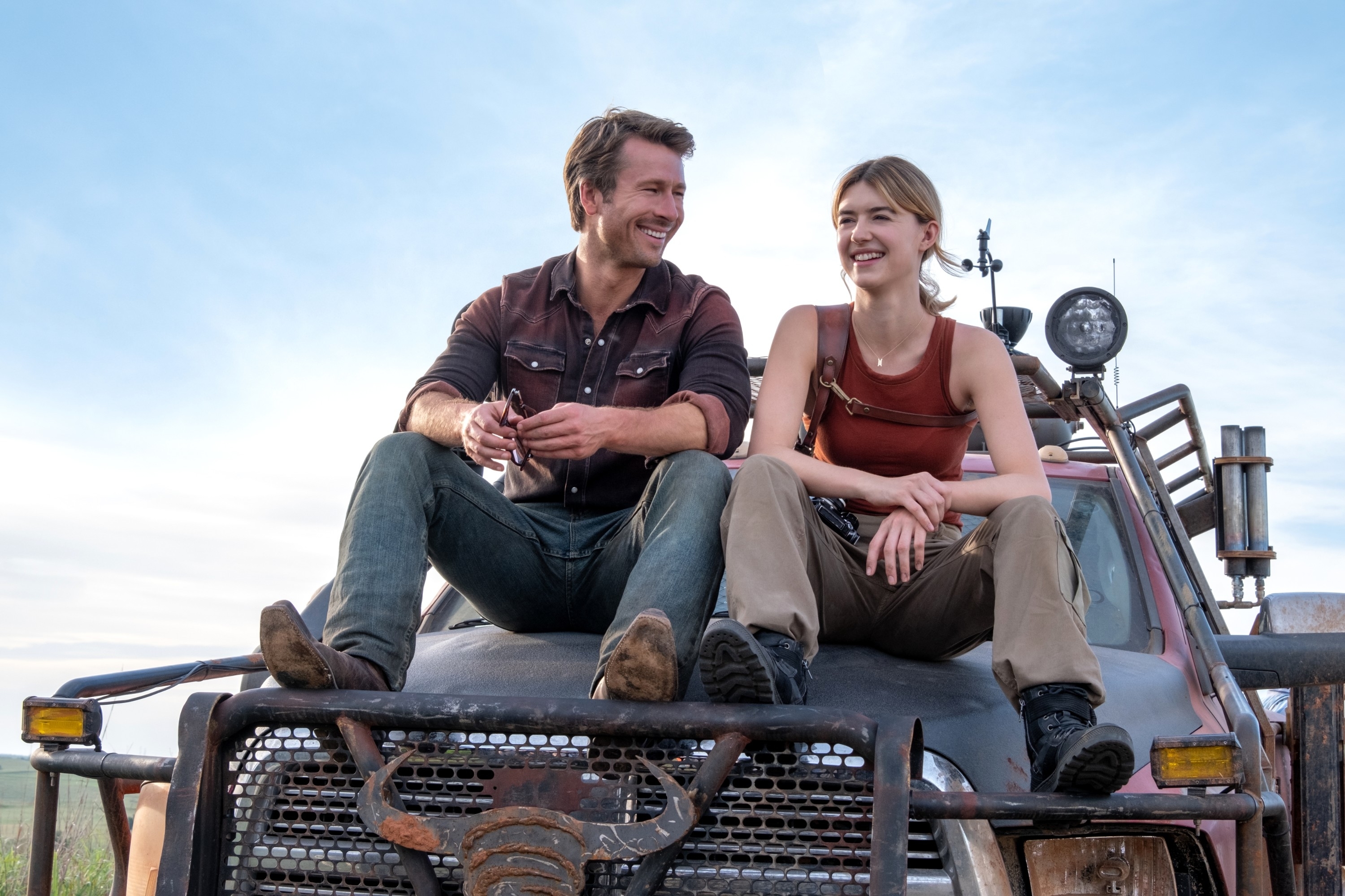 Glen Powell and Daisy Edgar-Jones sit on a rugged truck, smiling and chatting. Both wear casual, outdoorsy attire, with a picturesque landscape behind them
