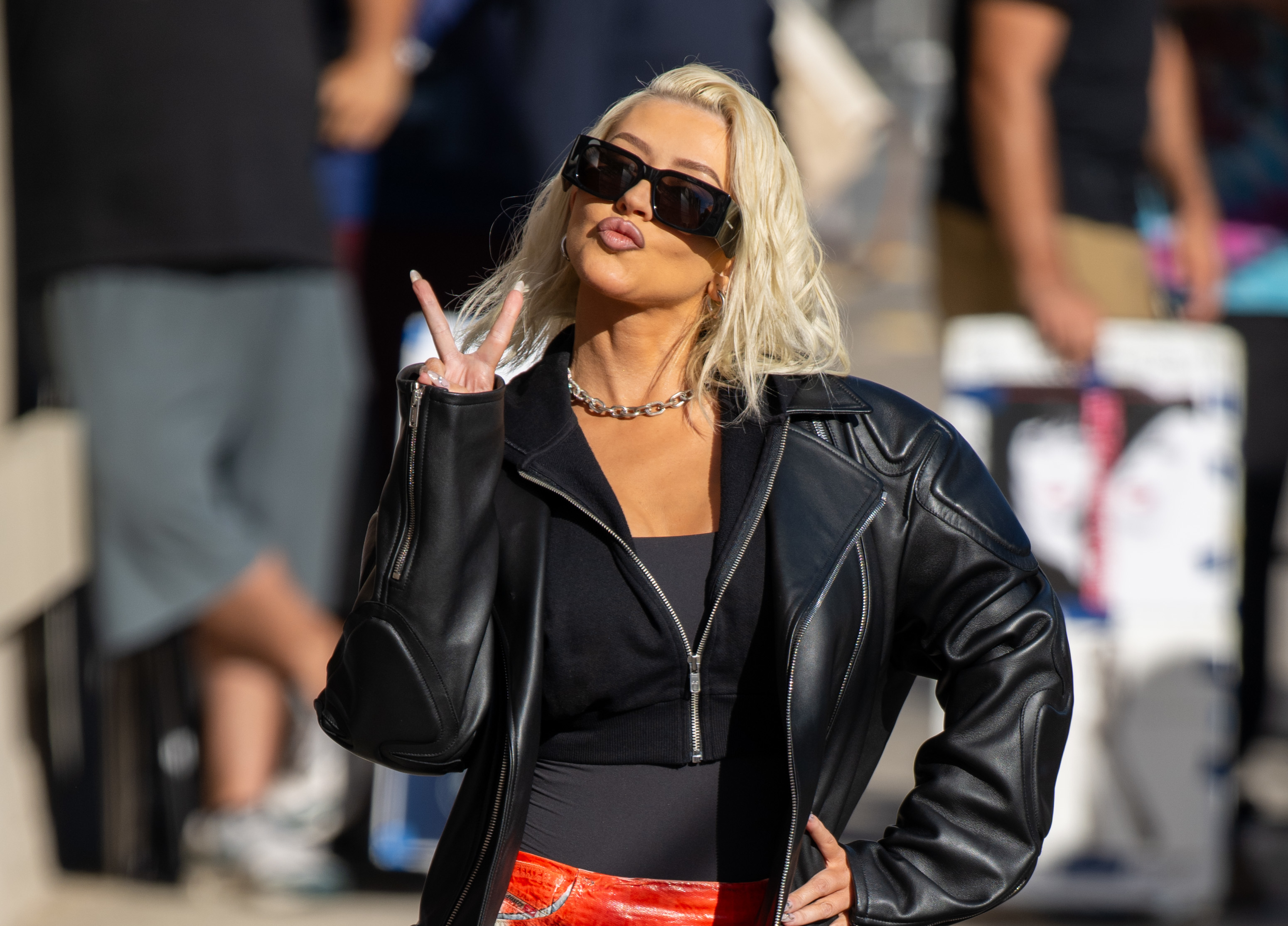 Christina Aguilera wearing a black leather jacket, black top, and red pants, makes a peace sign with one hand while posing for the camera