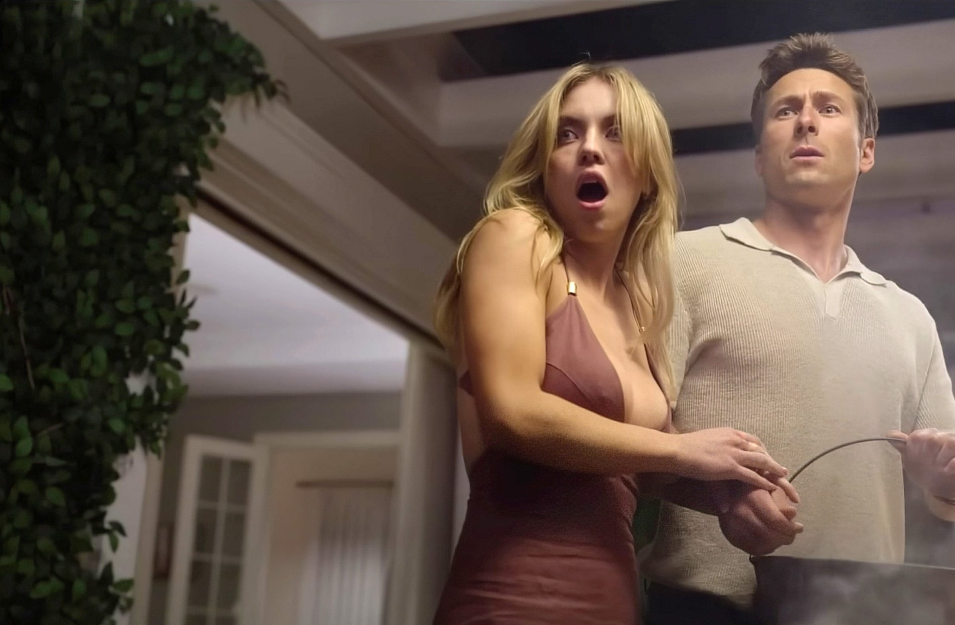 Sydney Sweeney and Glen Powell, both looking surprised, standing indoors in front of greenery and windows