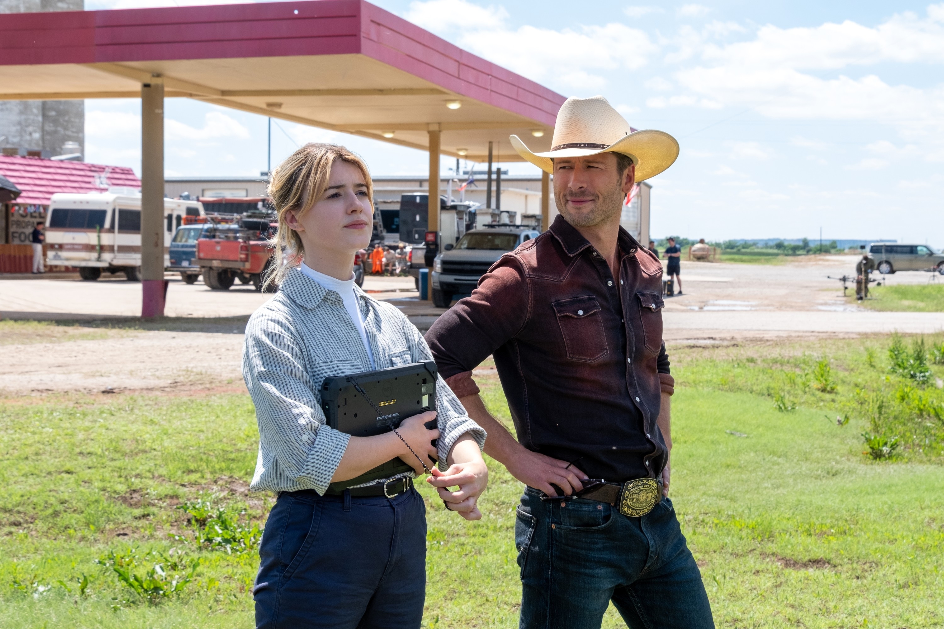 Daisy Edgar-Jones and Glen Powell stand outdoors near a gas station. Kristen holds a tablet while David wears a cowboy hat and belt buckle