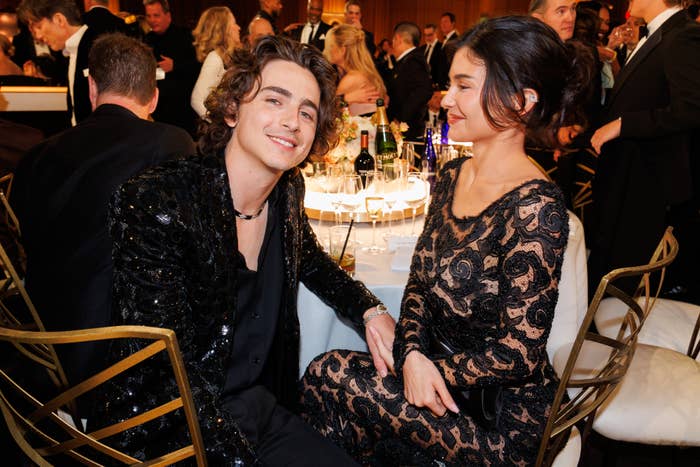 Timothée Chalamet and Kylie Jenner sitting at a table, smiling at an event. Chalamet is wearing a sequin jacket, while Gomez is in a lacy black dress
