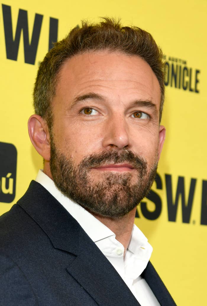 Ben Affleck at an event, wearing a suit jacket over a white dress shirt, looking toward the left with a yellow background