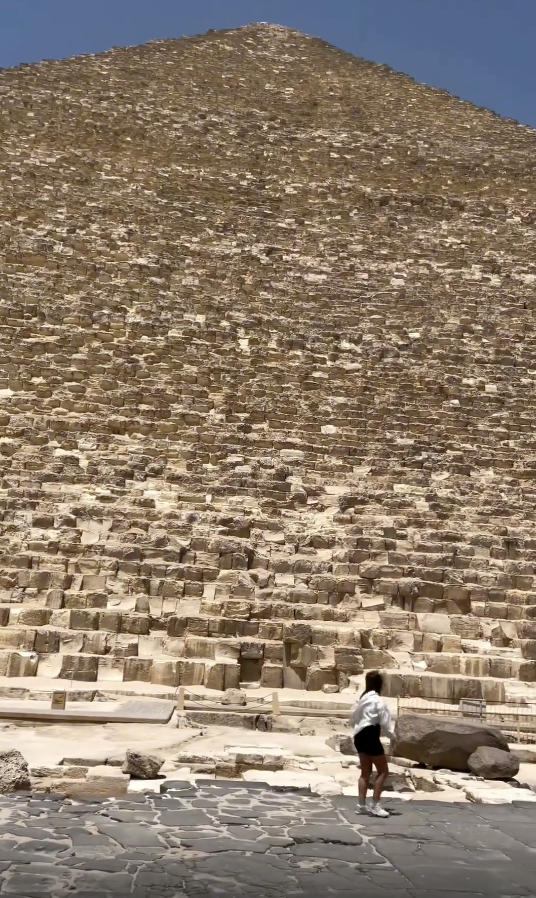 A person stands next to an ancient pyramid, looking up at the towering structure made of large stone blocks under a clear sky